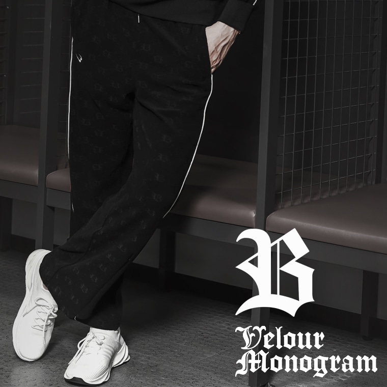 Velour Monogram。ホワイトのスニーカーを合わせ、ベンチにもたれて脚を組む男性モデルが着用する、モノグラム柄とホワイトパイピングが施された黒のベロアパンツ。